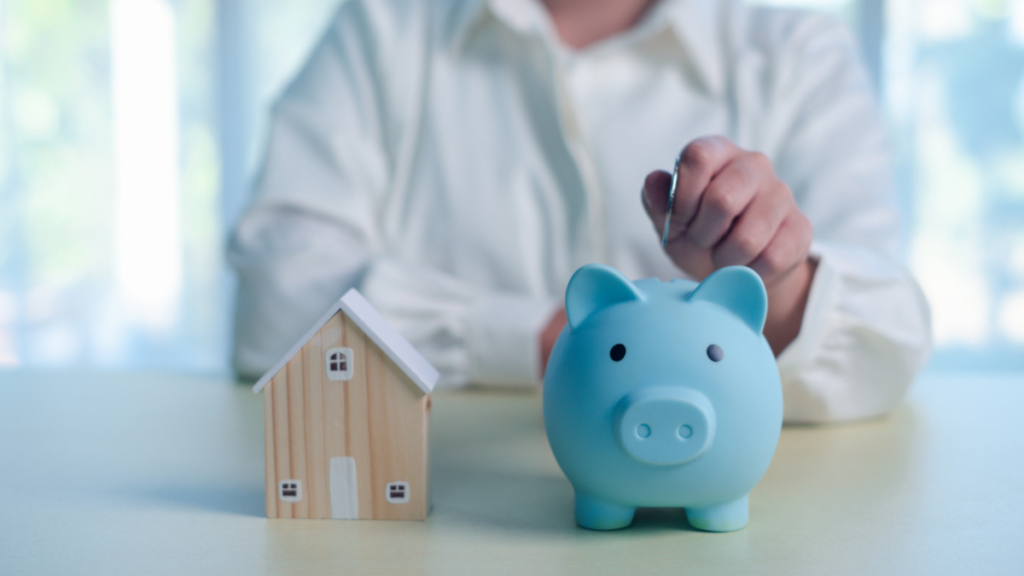 A blue piggy bank and a small wooden house model sit on a table, with a person in a white shirt and pen in the background, suggesting saving for a home or financial planning.